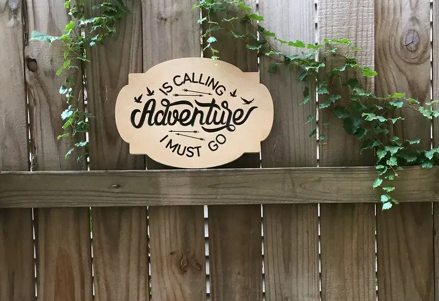 Finished wood sign reading Adventure Must Go displayed on a garden fence