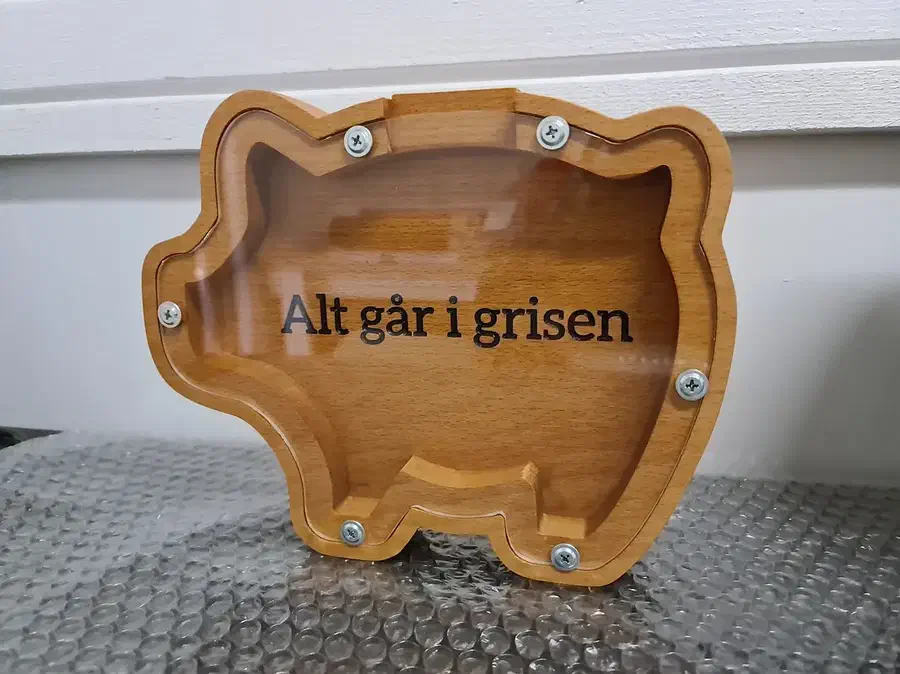 A wooden pig-shaped coin bank with a clear screwed-on front on a windowsill