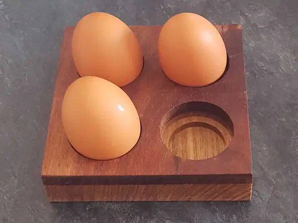 Three eggs beside an empty recess in a square holder made from contrasting timber layers