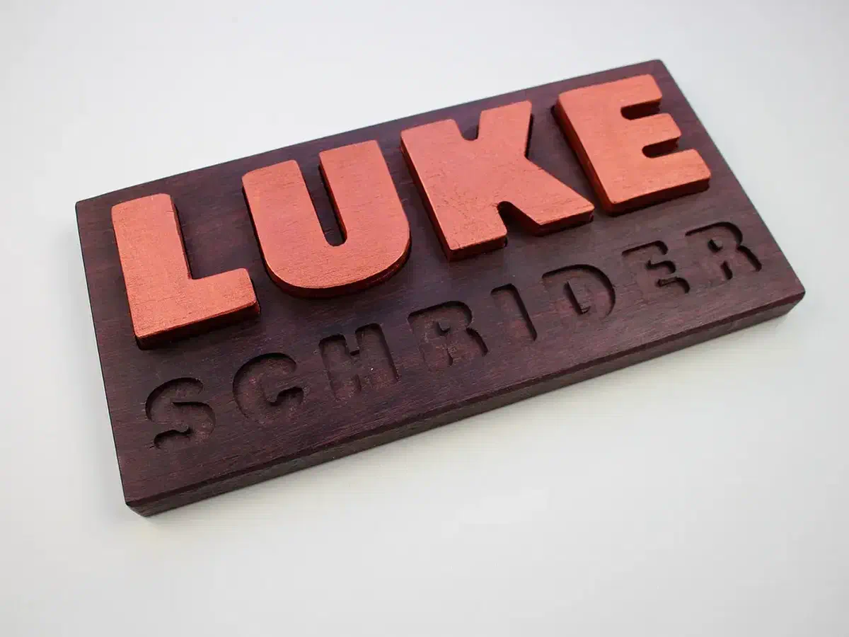Removable coral-colored LUKE letters on a dark wooden puzzle board with a carved surname