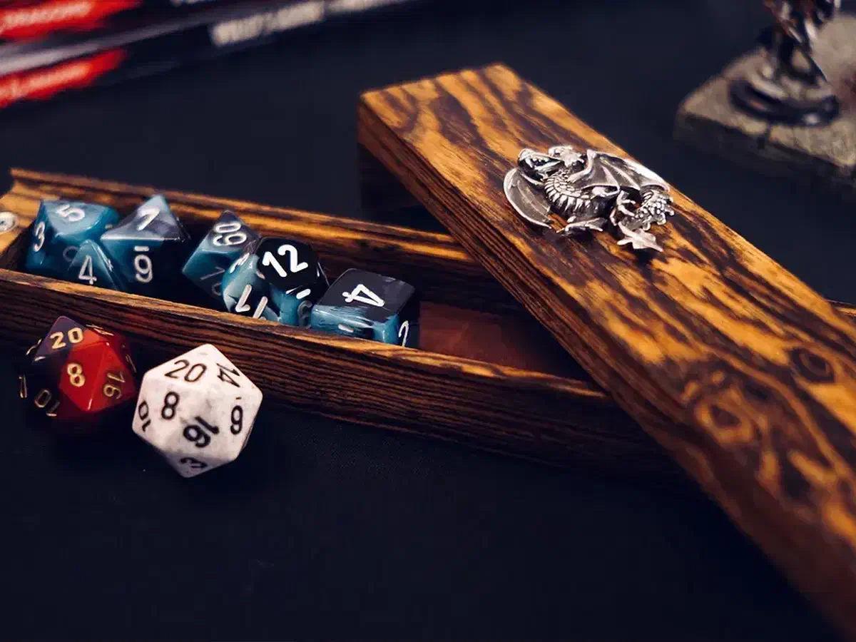 Open striped wooden dice vault holding blue dice beside its silver-dragon-decorated lid on a dark tabletop