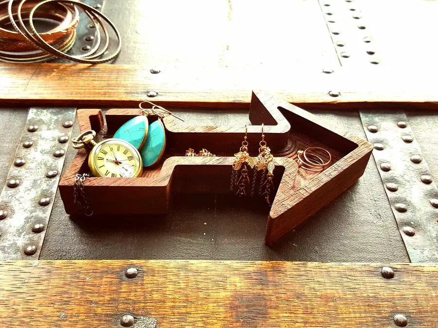 A walnut arrow-shaped dish holding a watch, earrings and rings on a wooden metal-trimmed chest