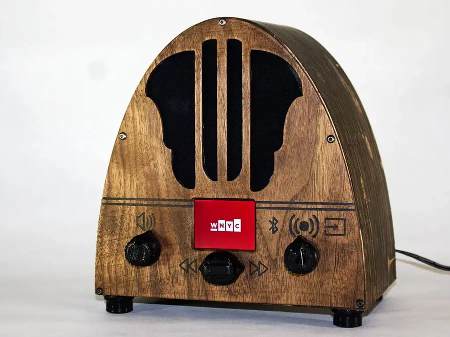 A finished arched wooden radio case with black speaker grille openings, a red display and three black controls on a white surface