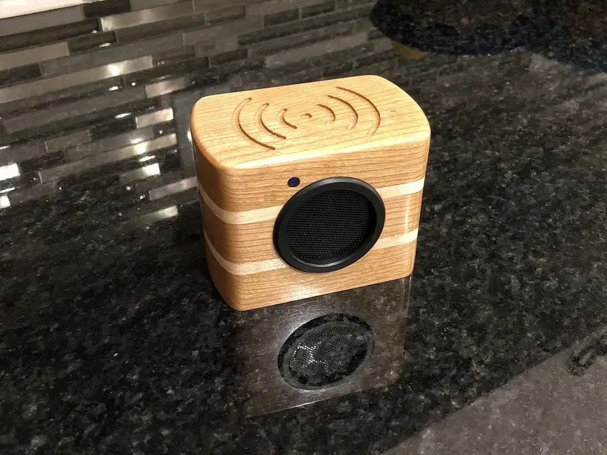 A small wooden speaker with a dark round grille and a pale stripe standing on a dark countertop