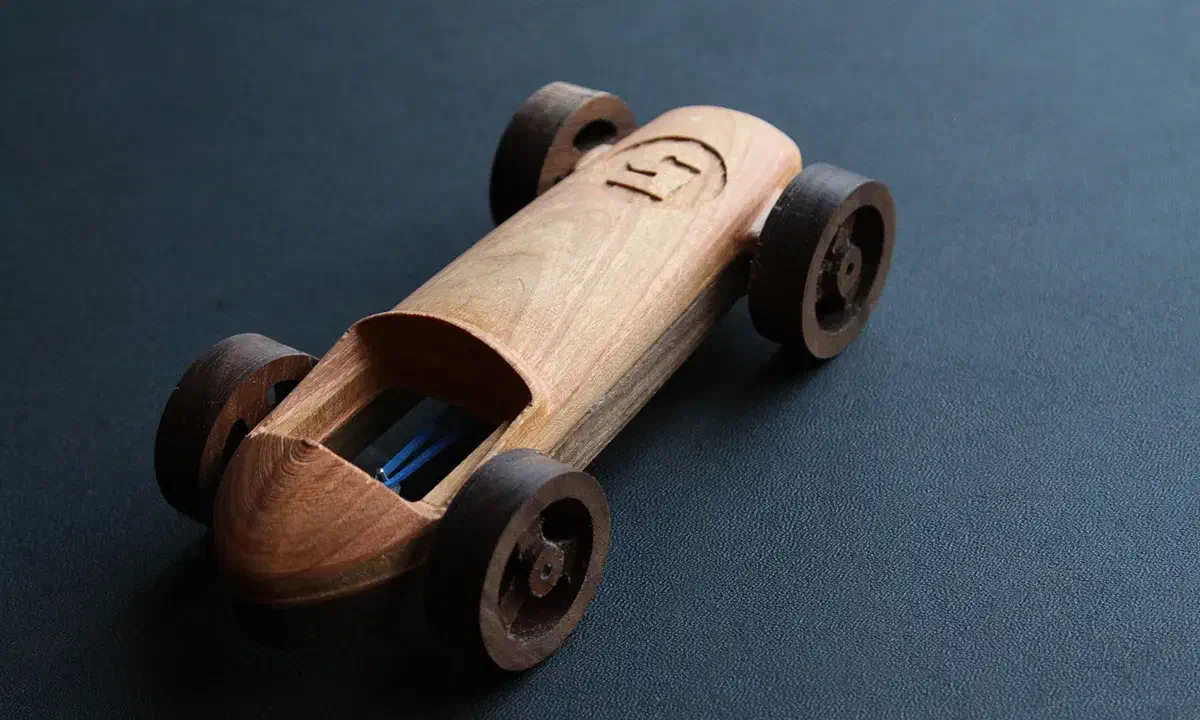 A brown wooden racing car with a carved rounded hood and large wooden spoke wheels on a dark surface